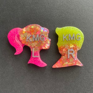 May include: Two resin silhouette figures of a girl and a boy with glitter embedded in the resin. The girl is pink and the boy is green. Both figures have the letters "KMG" and "L" or "R" on them.