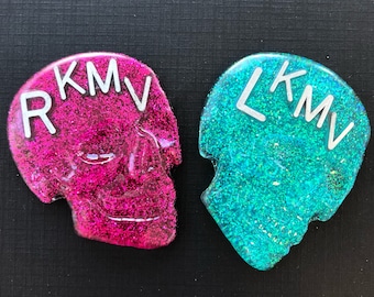 Può includere: Due spille in resina a forma di teschio con glitter incorporati nella resina. Una spilla è rosa con le lettere "RKM" in bianco, e l'altra è turchese con le lettere "LKM" in bianco.