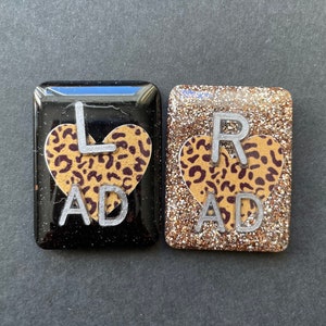 May include: Two small, square-shaped resin tiles with a leopard print heart design. The left tile is black with a silver "L AD" and the right tile is gold with a silver "R AD".
