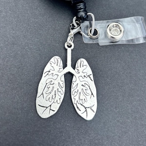 Puede incluir: Un dije plateado con forma de pulmones, sujeto a un portatarjetas retráctil. El portatarjetas tiene una cara blanca con el texto "The Love You Deserve" en una fuente cursiva. El fondo es gris oscuro.