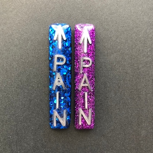 May include: Two glitter-covered rectangular stickers with the word "PAIN" written vertically in silver letters. The sticker on the left is blue and the sticker on the right is purple.