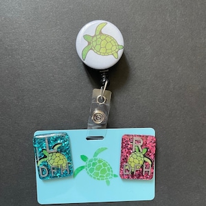 May include: A retractable badge reel with a light green turtle design on a white background. The badge holder has a light blue base with a turtle graphic and two glittery rectangles with letters and turtle graphics.