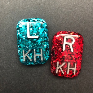 Puede incluir: Dos piezas rectangulares de resina rellenas de purpurina. Una es de color turquesa con la letra "L" y las letras "KH". La otra es roja con la letra "R" y las letras "KH".