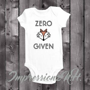 May include: White baby onesie with the text "ZERO GIVEN" in black outlined letters. A cartoon fox face with orange ears and a black nose is between the words.