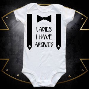 May include: White baby onesie with black suspenders and a bow tie. The onesie has the text "LADIES I HAVE ARRIVED" printed on it.