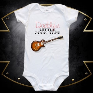 May include: White baby onesie with the text "Daddy's Little Rock Star" in black and red lettering. A brown electric guitar is pictured below the text.
