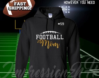 Op de afbeelding: Zwarte hoodie met een witte voetbalafbeelding en gouden glittertekst die "Football Mom" en het nummer "#59" op de voorkant zegt.