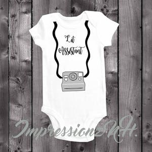 May include: White baby onesie with black text that reads "Lil' Assistant" and a black and gray illustration of an instant camera with a strap.