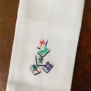 May include: White kitchen towel with embroidered mahjong tiles in red, green, blue, and white.