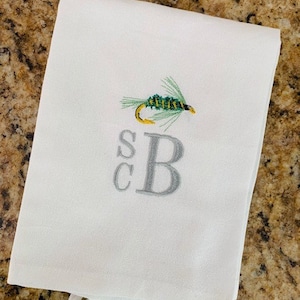 以下が含まれることがあります： グレーの刺繍された文字Bと、その上に文字SCが刺繍された白いキッチンタオル。文字Bの上に、緑と黄色のフライフィッシングルアーが刺繍されています。