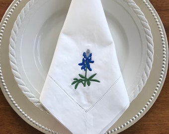 Peut inclure: Serviette en lin blanc avec un motif de fleur de bleuet brodé. La serviette est pliée en triangle et placée sur un chargeur argenté.