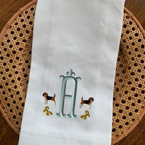 May include: White kitchen towel with a blue embroidered letter A and two brown and white dogs on either side of the letter. The dogs are facing each other and have their tails wagging.