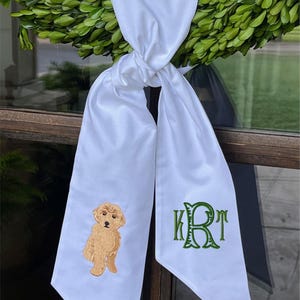 May include: White sash with a knotted top, featuring an embroidered golden doodle and green monogram. The sash is draped over a green wreath, creating a decorative accent. The design is simple and elegant.