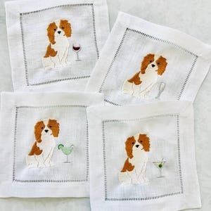 Könnte beinhalten: Ein Set aus vier quadratischen Cocktailservietten aus weißem Leinen, die jeweils einen gestickten braun-weißen Cavalier King Charles Spaniel neben einem anderen Cocktailglas zeigen, darunter Weinglas, Champagnerflöte, Margarita und Martini.