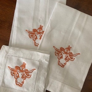 Könnte beinhalten: Weiße Leinentücher und ein quadratischer Untersetzer mit einem gestickten orangefarbenen Longhorn-Design. Die Handtücher haben einen einfachen Saum, und der Untersetzer hat eine dekorative genähte Bordüre. Das Design ist ein stilisierter Longhorn-Kopf mit Hut.