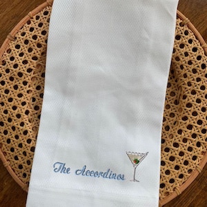 Può includere: Asciugamano da cucina bianco con la scritta ricamata "The Accordinos" in corsivo blu. Un bicchiere da martini con un'oliva è ricamato accanto al testo. L'asciugamano è piegato e posto su un vassoio di vimini intrecciato.