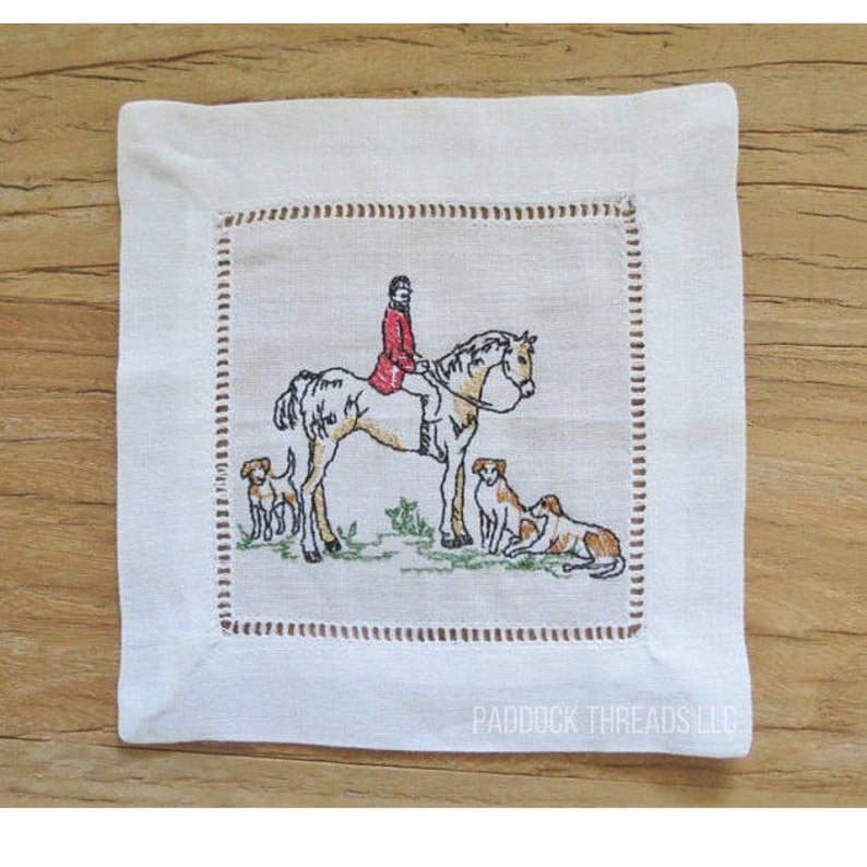 Puede incluir: Servilleta de c&oacute;ctel de lino blanco con una imagen bordada de una persona montando a caballo con dos perros. El texto "PADDOCK THREADS LLC" est&aacute; bordado en la esquina inferior derecha.