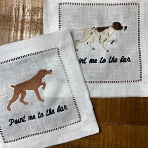 May include: Two white linen napkins with embroidered brown and white dogs and the text "Point me to the bar".