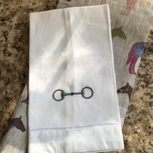 May include: White hand towel with an embroidered silver horse bit design.