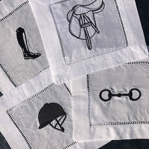 May include: Four white linen napkins with black embroidered designs. The designs include a riding boot, a saddle, a riding helmet, and a snaffle bit.