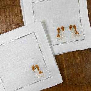 Könnte beinhalten: Zwei weiße Leinenservietten mit gestickten Cavalier King Charles Spaniel Hunden. Die Servietten haben einen weißen gestickten Rand.