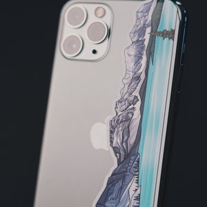 Puede incluir: Un iPhone plateado con una pegatina de una cordillera y un lago en la parte posterior.