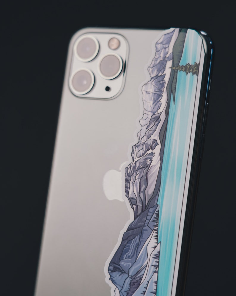 Puede incluir: Un iPhone plateado con una pegatina de una cordillera y un lago en la parte posterior.