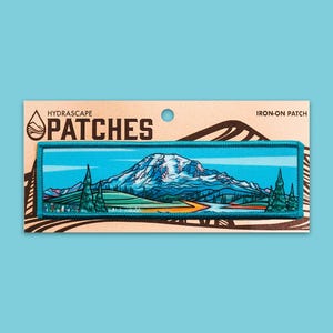May include: An iron-on patch featuring a mountain landscape with a river and trees. The patch is rectangular with a teal border and a mountain scene in shades of blue, white, and orange. The text "HYDRASCAPE PATCHES" is printed on the packaging.