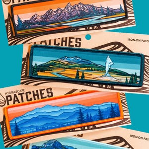 Puede incluir: Cuatro parches termoadhesivos con diseños de paisajes. Cada parche tiene forma rectangular con una ilustración detallada de montañas, árboles y agua. Los parches tienen un borde negro o de color y el texto "HYDRASCAPE PATCHES" es visible en el embalaje.