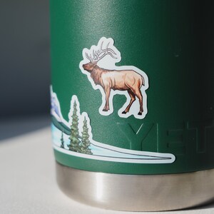 Puede incluir: Una botella de agua Yeti verde con una pegatina de un alce y una escena de monta&ntilde;a. La pegatina es blanca con detalles marrones y verdes.