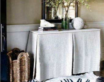 May include: A white linen table skirt with pleats covers a white table with a black mirror and a vase of greenery above it. The table is decorated with books and a white decorative object. A wicker basket is on the floor next to the table.