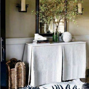 May include: A white linen table skirt with pleats covers a white table with a black mirror and a vase of greenery above it. The table is decorated with books and a white decorative object. A wicker basket is on the floor next to the table.