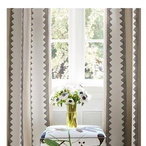 Pode incluir: Um otomano quadrado branco com um padrão floral verde, marrom e preto. O otomano está sentado em um tapete tecido na frente de uma janela com cortinas brancas.