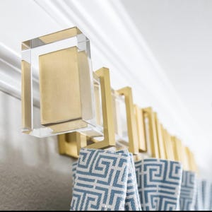 May include: A modern curtain rod with clear acrylic and gold-toned metal brackets. The rod holds blue and white patterned towels. The design is geometric and the overall aesthetic is contemporary.