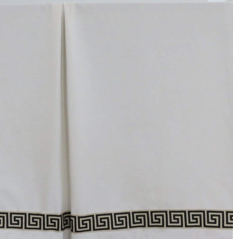May include: White fabric with a black and tan Greek key border design. The border is a repeating geometric pattern. The fabric is folded in the center, creating a vertical line. The overall look is clean and classic.