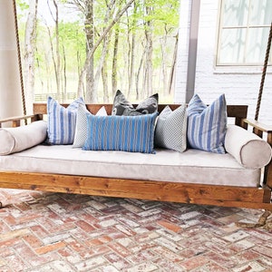 Puede incluir: Un columpio de porche de madera con un cojín de color claro y varias almohadas decorativas. Las almohadas presentan rayas azules y blancas, un patrón de cuadros y un patrón oscuro. El columpio está suspendido por cuerdas gruesas.