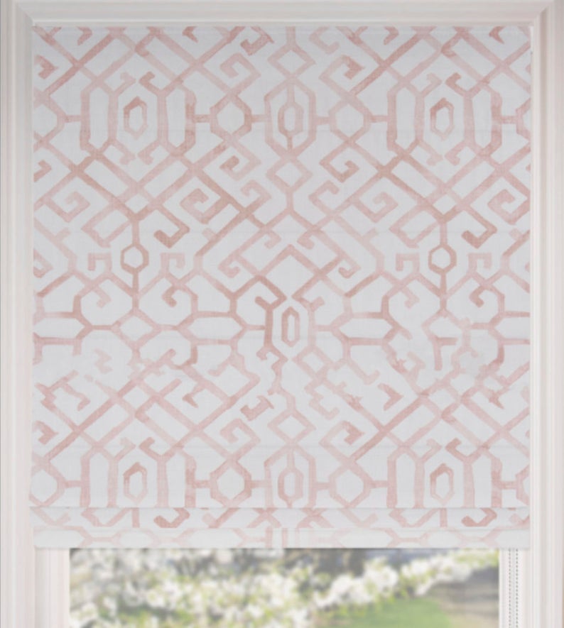 May include: A Roman shade with a geometric pattern in shades of blush pink and white. The fabric is a light color, and the design is a repeating series of interlocking shapes. The shade is partially lowered, revealing a window and a glimpse of the outside.