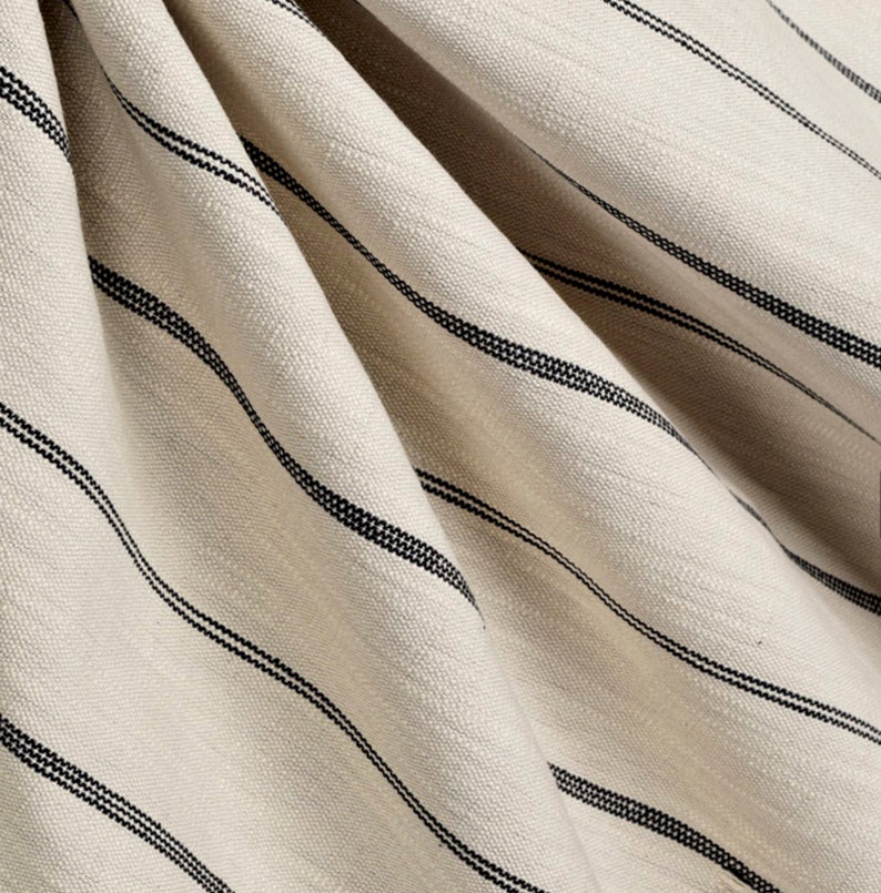 May include: Close-up of a cream-colored fabric with thin black stripes. The fabric is woven and has a subtle texture.