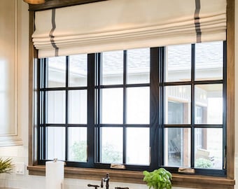 May include: A kitchen window with a black grid design, framed in wood. A cream-colored Roman shade with a gray stripe hangs above the window. The kitchen features a white sink, faucet, and countertop. Decorative lighting fixtures are mounted above the window.