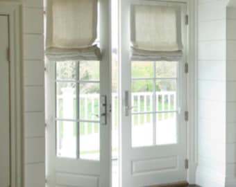 May include: White French doors with a view of a backyard through the glass panes. The doors have a white trim and are topped with a white roman shade.