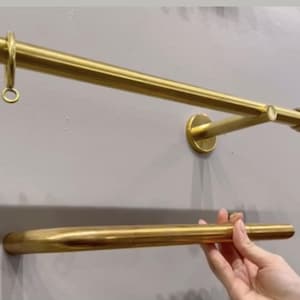 Pu&ograve; includere: Un set di bastoni per tende color oro contro un muro grigio. Il set comprende un bastone orizzontale con anelli, un bastone sporgente e un bastone inferiore tenuto in mano. I bastoni sono in metallo e hanno una finitura liscia.