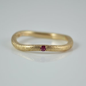 Könnte beinhalten: Ein dünner Goldring mit einem einzigen, kleinen, roten Edelstein in der Mitte.