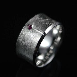 Könnte beinhalten: Ein breiter, gebürsteter Silberring mit einem kleinen, runden, roten Edelstein. Der Ring hat eine strukturierte Oberfläche und ein modernes Design, vor schwarzem Hintergrund. Der Edelstein ist in die Oberfläche des Rings eingesetzt.