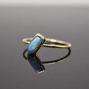 Könnte beinhalten: Ein goldener Ring mit einem rechteckigen Opalstein. Der Opal hat eine blaue und grüne Farbe mit einer goldenen Lünettenfassung. Der Ring ist vor einem dunklen Hintergrund platziert, mit einer Reflexion des Rings.