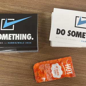 May include: A stack of black and white square stickers with a blue checkmark and the text "DO SOMETHING. MICHELLE OBAMA FOR HARRIS/WALZ 2024". There is also a stack of white square stickers with the same design and text. A small orange packet of hot sauce is in the foreground.