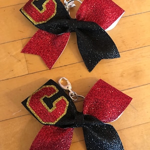 May include: Two cheerleading bows with a black center and red glitter accents. Each bow features a gold-outlined red letter "G" and a silver keychain clip. The bows are on a light brown wooden surface.