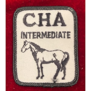 May include: A white patch with black text that reads "CHA INTERMEDIATE". Below the text is a black and white illustration of a horse. The patch has a black border and is set against a red background.