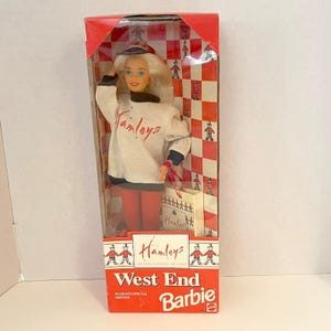 May include: A West End Barbie doll in a red and white box. The doll has blonde hair and is wearing a white sweatshirt with "Hamleys" in red script, red pants, and a hat. The box reads "Hamleys West End Barbie".
