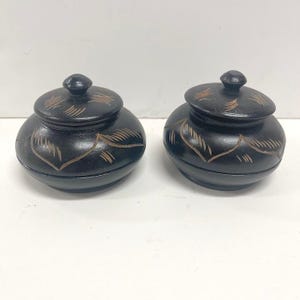 May include: Two small, round, black lidded containers with a smooth finish. Each container features a matching lid with a small knob handle. The exterior surfaces of both the lids and the bases are decorated with etched, light-colored leaf patterns.