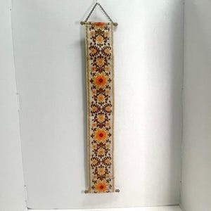 May include: A long, rectangular fabric wall hanging featuring a repeating floral pattern in shades of orange, yellow, and brown on a cream background. The textile is suspended from a thin metal chain attached to a horizontal rod at the top.